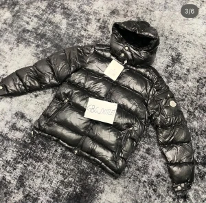 Svart Moncler pufferjacka med huva - Säljer en svart pufferjacka från Moncler med glansig finish och huva. Jackan har två stora fickor med dragkedja, Moncler-logga på ärmen och quiltad design. Perfekt för dig som vill ha en snygg och varm jacka med streetkänsla.