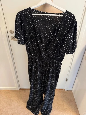 Svart prickig långklänning från SHEIN - Svart jumbsuit från SHEIN med vita prickar, v-ringad omlottfront och korta ärmar. Jumpsuit har resår i midjan för en snygg passform och är tillverkad i ett lätt, mjukt material som känns skönt mot huden.