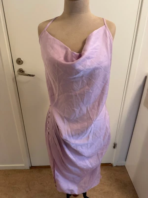 Ljuslila satinklänning Missguided Plus - Superfin ljuslila satinklänning från Missguided Plus med smala axelband, draperad halsringning och knappar längs sidan. Klänningen har en elegant, figurnära passform och slutar strax under knäet. Perfekt för dig som vill sticka ut med en mjuk pastellfärg.