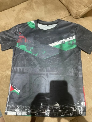 Svart t-shirt med Palestina-tryck - Unik svart t-shirt med tryck av stadssilhuett och palestinska flaggan i rött, grönt och vitt. T-shirten har rund hals och korta ärmar, med coola grafiska detaljer över hela framsidan. Perfekt för dig som vill sticka ut med ett statement-plagg.