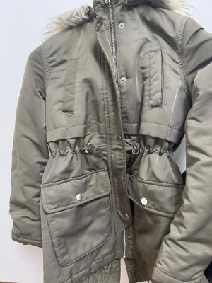 Olivgrön parkas från L.O.G.G. med huva - Parkas i olivgrönt från L.O.G.G. med fluffig huva, stora fickor med tryckknappar och snörning i midjan. Jackan har dragkedja och knappstängning framtill, samt långa ärmar. Perfekt för kalla dagar med sin fodrade insida och praktiska detaljer.