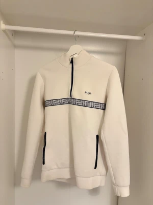 Vit half zip sweatshirt från BOSS - Vit sweatshirt från BOSS med half zip och hög krage. Svartvit BOSS-logoband går tvärs över bröstet och svarta detaljer vid fickorna. Långärmad och i mjukt bomullsmaterial, perfekt för chill dagar. Snygg och stilren design med sportig vibe. Ny pris 1499kr