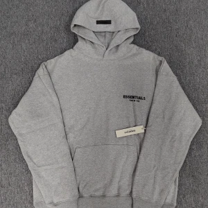Grå hoodie Essentials Fear of God - Säljer en grå hoodie från Essentials Fear of God med svart logotyp på bröstet. Tröjan har huva, lång ärm och en stor magficka framtill. Perfekt för dig som gillar streetwear och enkel stil. Mjuk bomullskänsla och relaxed passform. Helt nytt. fraktar inom 24 timmar. för mer info skriv t mig.