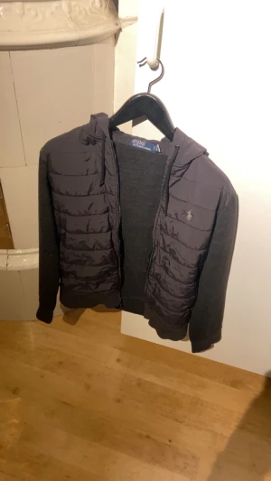 Svart cardigan från Polo Ralph Lauren - Svart pufferjacka från Polo Ralph Lauren med huva och dragkedja. Jackan har quiltade paneler framtill, mjuka stickade ärmar och en broderad logga på bröstet. Perfekt för dig som vill ha en stilren och bekväm jacka med cool streetkänsla.