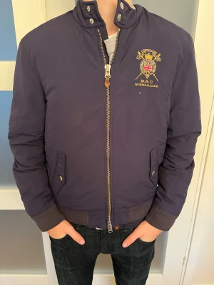 Morris Rowing Club mörkblå bomberjacka - Mörkblå bomberjacka från Morris Rowing Club med broderad klubbemblem på bröstet. Jackan har ribbade muddar, ståkrage med tryckknappar och dragkedja framtill. Två fickor med knappstängning och snygga detaljer i guld. Perfekt för dig som gillar klassisk stil.