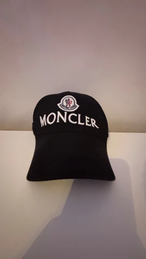 Svart Moncler keps med broderad logga - Snygg svart keps från Moncler med stor vit broderad logga framtill och klassisk Moncler-emblem ovanför. Justerbar baktill för perfekt passform. Materialet är mjukt och bekvämt, perfekt för att lyfta din streetwear-look.