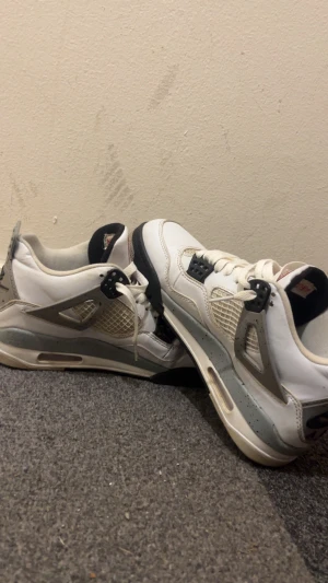 Nike Air Jordan 4 vit/grå sneakers - Nike Air Jordan 4 sneakers i vit och grå med svarta detaljer. Skorna har meshpaneler på sidorna, synlig Air-sula och klassisk snörning. Grå häl med svart Nike Air-logga och stänkmönster. Tungan är svart med Air Jordan-märke i rött. Perfekt för dig som gillar retrostil.