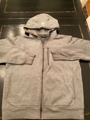 Grå hoodie från Sail Racing - Snygg grå zip-hoodie från Sail Racing med broderad logga på huvan och stor text på ryggen. Mjuk insida, dragkedja framtill. Dragkedjan för bröstfickan saknar ”handtag” men fickar går fortfarande att dra upp och stänga. Finns ett litet stygn på baksidan om man zoomar lite kan man se