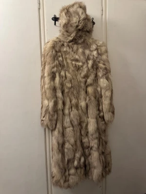 Beige lång pälsjacka med huva -  Säljer en riktigt fluffig och lång pälsjacka i beige ton med snygga mörkare inslag. Jackan har en stor huva och långa ärmar, perfekt för dig som vill sticka ut och hålla dig varm. Den har en loose passform och är supermysig att bära. Äkta blårävspäls. Nypris 20000:-