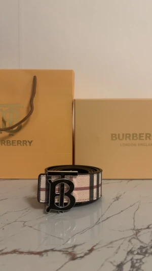 Burberry bälte - Säljer ett Burberry bälte med klassiskt randigt mönster i beige, svart och röd. Bältet har ett stort metallspänne med Burberrys ikoniska B-logga. Ena sidan är i textil med mönster, andra sidan är svart skinn med präglad rutig design och ryttare.