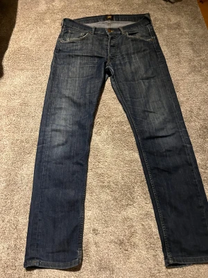 Mörkblå jeans från Lee - Snygga mörkblå jeans från Lee med klassisk femficksdesign och raka ben. Jeansen har subtila slitningar och kontrastsömmar, vilket ger en cool och avslappnad look. Perfekta för dig som gillar tidlös stil och vill ha ett par jeans som funkar till det mesta.