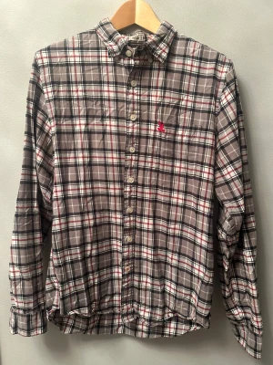 Rutig skjorta CC Double O L - Snygg rutig skjorta från CC Double O i storlek L. Skjortan har en klassisk krage, knappar hela vägen och ett broderat rött fågelmotiv på bröstet. Färgerna är beige, svart, vit och röd. Tillverkad i mjuk bomull, perfekt för dig som gillar avslappnad stil.