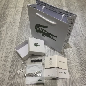 Lacoste silverfärgat armband - Snyggt armband från Lacoste i silverfärgat rostfritt stål med stilrena, rektangulära länkar och diskret Lacoste-logga. Kommer med originalförpackning, tygpåse och verktyg för justering. Perfekt accessoar för dig som gillar minimalistisk och modern stil.