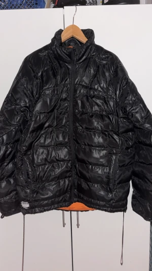 Svart pufferjacka med orange foder - Snygg svart pufferjacka från Svea med glansig finish och orange foder. Jackan har dragkedja framtill, två fickor med dragkedja och meshficka på insidan. Perfekt för kalla dagar och har en modern, sportig vibe.