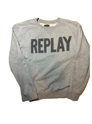 Grå sweatshirt från Replay - Snygg grå sweatshirt från Replay med stor svart logga över bröstet. Tröjan har rund halsringning, långa ärmar och ribbade muddar vid ärmslut och nederkant. Perfekt för en avslappnad och trendig stil.