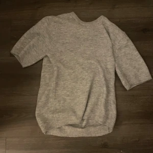 Grå stickad tröja med trekvartsärm - Mysig grå stickad tröja med trekvartsärmar och rund halsringning. Tröjan har en avslappnad passform och ribbade muddar vid ärmslut och nederkant. Perfekt för lager-på-lager-stil under kyliga dagar.