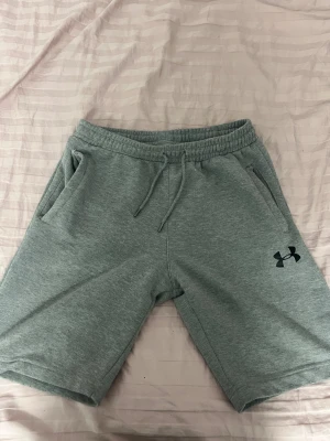 Grå shorts från Under Armour - Snygga grå shorts från Under Armour med elastisk midja och snörning. Mjukt material och två sidofickor, perfekt för chill eller träning. Klassisk logga på ena benet och enkel, stilren design.