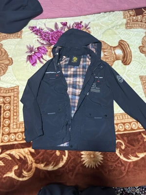 Svart vindjacka Leduntino M - Svart vindjacka från Leduntino i storlek M. Jackan har huva med snörning, två stora fickor framtill och coolt tryck på bröstet. Insidan är fodrad med rutig beige och blå textil. Perfekt för dig som gillar street och vill ha en snygg jacka med detaljer.