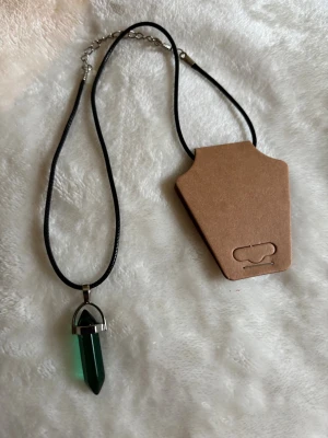 Halsband med grön kristallhänge - Snyggt halsband med svart läderimitation och ett spetsigt grönfärgat kristallhänge i silverfärgad infattning. Perfekt accessoar för dig som gillar kristaller och en stilren look.