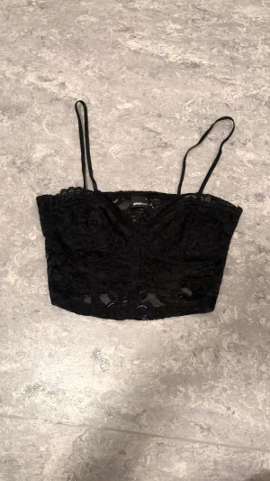 Svart spetsig korsettopp Gina Tricot - Säljer en svart korsettopp från Gina Tricot med tunna axelband och vacker spets över hela toppen. Croppad modell som ger en snygg siluett och passar perfekt till höga jeans eller kjol. Toppen har en feminin och trendig look.