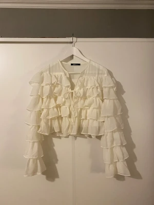 Volangtopp från gina - Vit topp med volang och långa ärmar. Genomginlig ovanför volangen men annars täckande. Knyte framtill och med liten krok/knapp en bit under knytet. Aldrig använd pga storlek