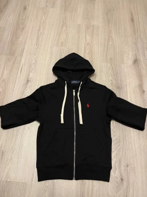 Svart hoodie från Polo Ralph Lauren - Svart zip-hoodie från Polo Ralph Lauren med klassisk röd logobrodyr på bröstet. Hoodien har vita snören, fickor fram och ribbade muddar. Tillverkad i mjuk bomullsmix som är skön mot huden. Perfekt för chill dagar och streetwear-stil.