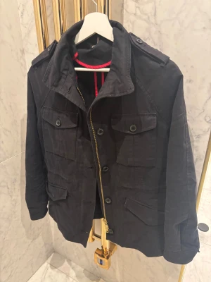 Jacka J.lindeberg  - Säljer en svart parkajacka från J.Lindeberg med fyra stora fickor framtill, axelklaffar och snygga detaljer i guld på dragkedjan. Skön passform och storlek XS. Snyggt och skönt material. Riktigt bra skick och inga defekter hittade! Jackan har röd insida med svarta och röda detaljer och stängs med både knappar och dragkedja. Pris: 1599kr 