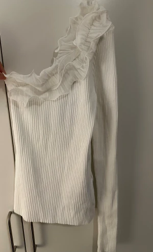 Vit oneshoulder topp med volang från Amisu - Säljer en vit ribbad oneshoulder topp från Amisu med lång ärm och stor volangdetalj i mesh längs halsringningen. Toppen har en tight passform och är perfekt för dig som vill sticka ut med en unik och trendig look.