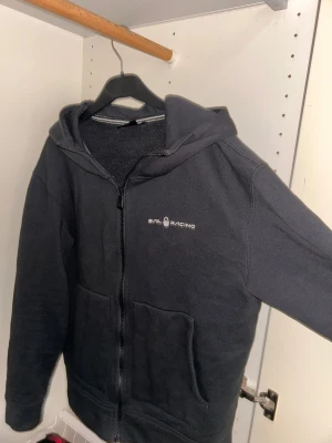 Svart hoodie från Sail Racing - Svart hoodie från Sail Racing med vit logga på bröstet och en grafisk detalj på huvan. Tröjan har dragkedja framtill, två stora fickor och är tillverkad i mjuk fleece. Perfekt för dig som gillar sportig och avslappnad stil.