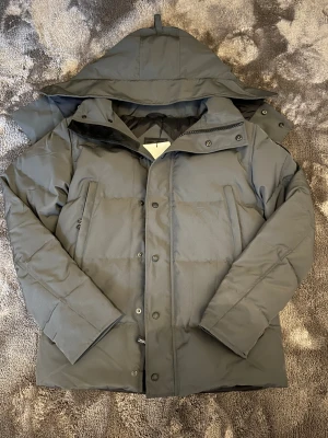 Canada goose jacka  - Säljer en grå dunjacka . Jackan har huva, dragkedja och knappar framtill samt två stora fickor.  Perfekt för kalla vinterdagar. Skriv vid eventuella frågor!