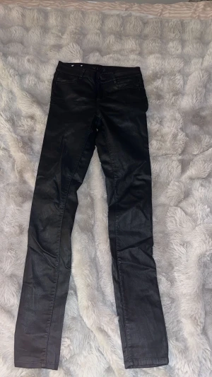 Svart coated jeans VILA - Snygga svarta coated jeans från VILA  med en glansig finish som ger en cool look. Modellen är skinny med normal passform och klassiska fickor både fram och bak. Perfekt för dig som gillar stilrena och trendiga byxor med lite edge. Dock ett håll vid bälteshållaren bak 