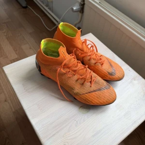 Orangea fotbollsskor med snörning - Säljer ett par grymt snygga orangea fotbollsskor med högt skaft och snörning. Skorna har en stickad ovandel och svarta detaljer framtill samt på hälen. Insidan är neongul för extra pop. Perfekta för dig som vill sticka ut på planen!