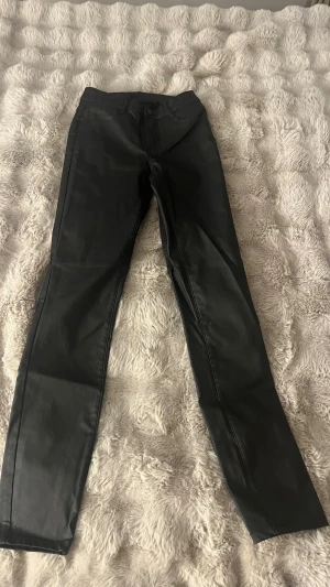 Svarta coated byxor från Vila - Svarta coated byxor från Vila i storlek S. Byxorna har en rak passform med klassiska fickor bak och bälteshällor. Materialet har en snygg, lätt glansig finish som ger en cool look. Perfekta för dig som vill ha ett par stilrena och trendiga byxor.