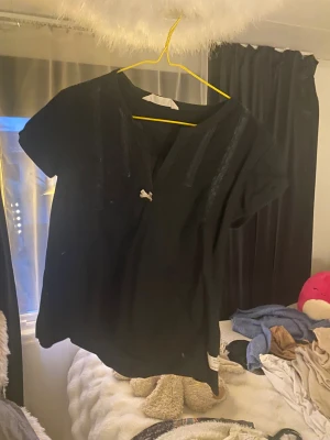 Svart blus med spets och rosett - Svart blus med kort ärm och v-ringning, dekorerad med smala spetsdetaljer längs bröstet och en liten vit rosett framtill. Perfekt för dig som gillar en söt och stilren look.