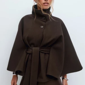 Mörkbrun kappa med bälte och hög krage - Cape jacket ginatricot  - Helt ny i XS/S 🩷 Snygg mörkbrun kappa med breda ärmar, hög krage och knappar framtill. Jackan har ett matchande bälte i midjan som ger en cool siluett. Materialet är mjukt och känns lyxigt, perfekt för dig som gillar stilrena och trendiga plagg.