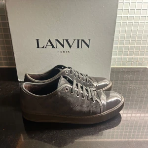 Grå sneakers från Lanvin med lackad tå - Snygga grå sneakers från Lanvin med en unik design där tån är i blankt lackat material och resten av skon i mjuk mocka. Skorna har rund tå, snörning och platt sula. Insidan är fodrad med skinn för extra komfort. Perfekt för dig som gillar exklusiva detaljer.