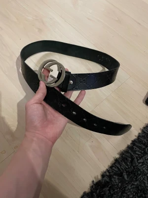 Svart bälte med GG-spänne - Snyggt svart bälte med präglade GG-loggor längs hela remmen och ett stort, rundat metallspänne med GG-design. Bältet är gjort i äkta skinn och har en klassisk, stilren look som lyfter vilken outfit som helst.