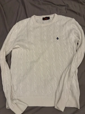 Vit kabelstickad tröja Morris M - Snygg vit kabelstickad tröja från Morris i storlek M finns en liten defekt i armhålan men det är lättfixat. Tillverkad av 100% pima bomull, med rund hals och ribbade muddar. Tröjan har ett mörkblått broderat emblem på bröstet och klassisk kabelstickad struktur som ger en clean och fräsch look.