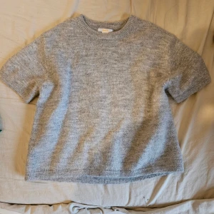 Grå stickad kortärmad tröja - Mjuk och skön grå stickad tröja från Soft Goat i kashmir. Tröjan har rund hals och korta ärmar, perfekt för lager-på-lager under kyliga dagar. Enkel och stilren design som funkar till många olika looks.