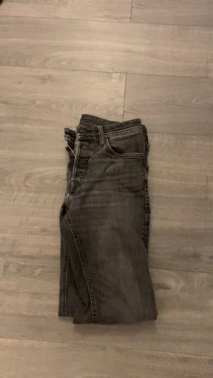 Grå archive jeans  - Snygga grå jeans från JACK AND JONES med slim/regular passform och klassisk femficksdesign. Jeansen har en mörkgrå tvätt med lätt slitna detaljer och raka bakfickor med broderad söm. Perfekt för dig som gillar en smal siluett och stilren look. Passar dig perfekt om du gillar archive stilen/grisch. Storleken är 29/32 