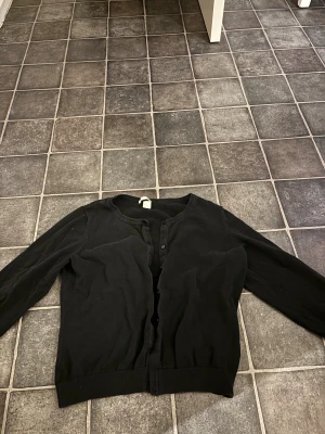 Svart kofta i bomull, storlek L - Svart kofta från H&M i storlek L sitter som S, Klassisk modell med rund hals, långa ärmar och knappar framtill. Tillverkad i mjuk bomull som är skön mot huden. Perfekt att slänga på sig när det är lite kyligt ute.