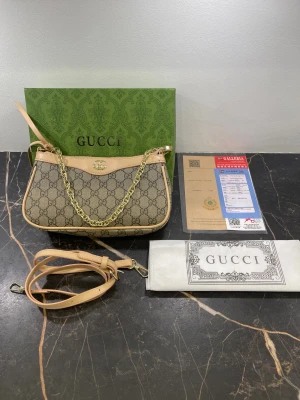 Beige handväska från Gucci med kedja - Snygg handväska från Gucci i beige och brunt med klassiskt GG-mönster. Väskan har guldfärgad kedja, ljusbeiga skinn-detaljer och dragkedja upptill. Inuti finns ett fack med präglad Gucci-logga. Perfekt storlek för det viktigaste och med justerbar axelrem.