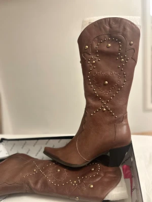 Bruna boots från Alfio Raldo - Snygga bruna cowboy boots i skinn från Alfio Raldo med spetsig tå och låg klack. Dekorerade med guldfärgade nitar i ett hjärtmönster på skaftet och har dragkedja på insidan. Perfekta för dig som vill sticka ut med western-vibe och coola detaljer. Svart päls i insidan.