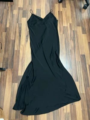 Svart långklänning från ASOS DESIGN - Svart långklänning med smala axelband från ASOS DESIGN. Klänningen har en elegant v-ringad hals och är figurnära upptill med ett mjukt fall nedtill. Materialet är lätt och känns skönt mot huden, perfekt för dig som gillar stilrena och minimalistiska looks.