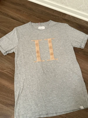 Grå t-shirt Les Deux med beige II - Snygg grå t-shirt från Les Deux med stort beige II-tryck i frotté på bröstet. Klassisk rund hals och korta ärmar. Mjuk och skön bomullskvalitet, perfekt för dig som gillar stilrena detaljer och enkel design.