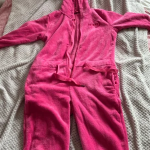 Rosa frotté jumpsuit med huva - Supermysig rosa jumpsuit i mjuk frotté med huva, dragkedja framtill och snörning i midjan. Perfekt för chill hemma eller sleepovers. Fickor på sidorna och broderad text på bröstet.