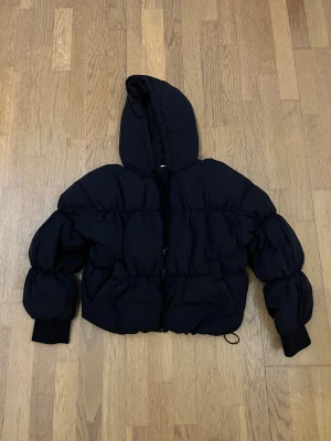 Svart pufferjacka från Divided - Svart pufferjacka från Divided i storlek S. Jackan har en stor huva, dragkedja framtill och puffiga, quiltade sektioner på både kropp och ärmar. Muddar vid ärmslut och justerbar nederkant med snörning. Perfekt för kalla dagar.
