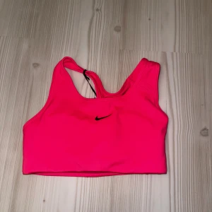 Nike rosa sport-bh Swoosh - Nike Swoosh sport-bh i starkt rosa med svart logga framtill. Bh:n har brottarrygg och är tillverkad i ett stretchigt, snabbtorkande syntetmaterial med Dri-FIT-teknologi. Perfekt för träning och ger medium support. Säljs med originaltaggar kvar.