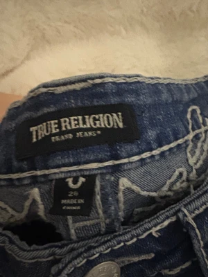 True Religion blå bootcut jeans - Säljer ett par blå jeans från True Religion med snygga vita kontrastsömmar och broderade detaljer på bakfickorna. Modellen är lågmidjad och har bootcut-ben. Klassisk knappstängning och coola ficklock bak. Perfekt för dig som gillar statement-jeans!