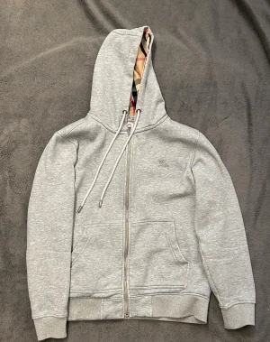 Grå Burberry hoodie med dragkedja - Snygg grå hoodie från Burberry i storlek S med klassisk dragkedja och två fickor framtill. Insidan av huvan har det ikoniska Burberry-rutiga mönstret i beige, svart och rött. Materialet är mjukt och bekvämt, perfekt för chill dagar. Broderad Burberry-logga på bröstet.